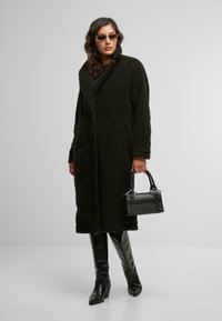 Cappotto lungo nero, texturizzato, con colletto a scialle, tasche frontali e vestibilità rilassata. Indossato con stivali neri lucidi fino al ginocchio e una piccola borsa.