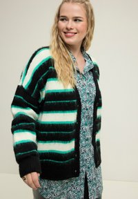 Gestreifter Strickcardigan in Schwarz, Weiß und Türkis mit weicher Textur und Knöpfen an der Vorderseite, über einem mit Paisleymuster bedruckten Hemd getragen.