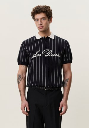 Jeune homme aux cheveux bouclés portant un polo rayé noir et blanc avec le texte "Les Deux" et un pantalon noir, debout devant un fond clair.
