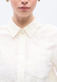 Biela kvetinová čipkovaná blúzka s golierovým výstrihom, zapínaním na gombíky a náprsným vreckom. Textúra je jemná a priehľadná.