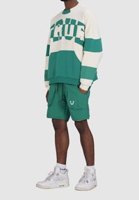 Zielono-biała bluza w paski z nadrukiem "TRUE", w połączeniu z zielonymi szortami z przednimi kieszeniami i logo. Całość dopełniają białe trampki.