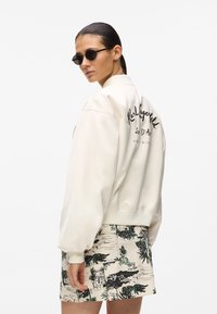 Veste bomber crème avec texte noir brodé dans le dos, associée à une jupe beige à motifs ornée de dessins floraux verts et noirs.