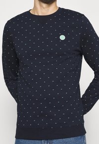 Sudadera azul oscuro con pequeños acentos verdes con patrón, cuello redondo, puños y dobladillo acanalados, y un parche de logo circular en el pecho.