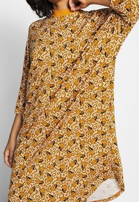 Dlouhé volné šaty vyrobené z měkké tkaniny v hořčicové žluté barvě s hravým vzorem černých a oranžových tvarů, s 3/4 rukávy.