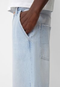 Hellblaue Jeans mit lockerer Passform, ausgestattet mit einer vorderen Tasche und einer aufgesetzten Tasche an der Seite. Der Stoff hat eine glatte Textur.