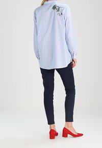 Camicia a righe blu chiaro con un dettaglio di ricamo floreale verde sulla schiena, abbinata a pantaloni blu navy e tacchi in suede rossi.