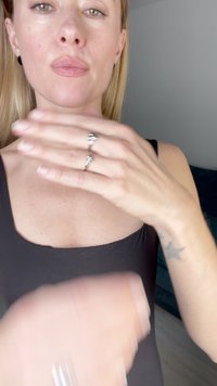 Modelo vistiendo una blusa negra con un escote redondeado, mostrando un anillo de plata en su mano. El fondo presenta una superficie texturizada de color azul oscuro.