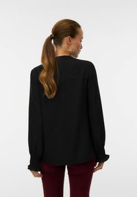 Donna con una lunga coda di cavallo che indossa una blusa nera a maniche lunghe e pantaloni bordeaux, in piedi di spalle contro uno sfondo semplice.