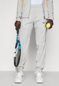 Lichtgrijze sportbroeken met elastische tailleband, taps toegesneden pijpen en met rits afsluitbare zakken. Houdt een tennisracket in de ene hand en een tennisbal in de andere.