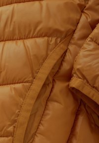 Brun vadderad jacka med detaljer som visar en slät, blank väv med kviltad textur, kontrasterande matt kant vid kanterna.