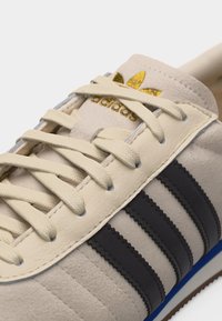Beige Adidas-tennari, jossa mustat raidat, beiget kengännauhat ja kultainen Adidas-logo kielen päällä.