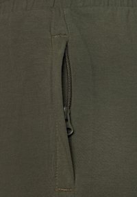 Olivgrüne Jogginghose mit glatter Textur, ausgestattet mit einer seitlichen Reißverschlusstasche und verstärktem Nähdetail. Der Reißverschluss ist aus schwarzem Metall.