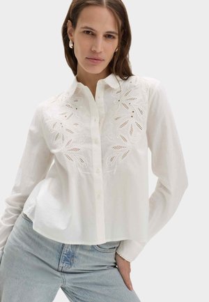 Kvinde iført en hvid broderet knap-op bluse og lyseblå jeans, poserende med den ene hånd i lommen mod en ensfarvet baggrund.