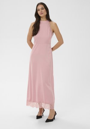 Kobieta stoi z rękami za plecami, ubraną w różową, bezrękawnikową sukienkę maxi z koronkowym dołem oraz czarne szpilki na prostym tle.
