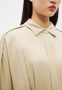 Žena oblečená v béžovej košeli na gombíky s ostrým golierom, zobrazená od ramien po bradu na bielom pozadí.