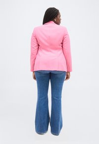 Γυναίκα με πλεξούδες, που φορά ροζ σακάκι, μπλε τζιν καμπάνα και λευκά παπούτσια, στέκεται πλάτη σε απλό φόντο.