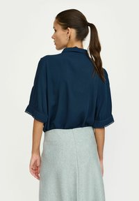 Navyblauwe blouse met korte poffende mouwen, voorzien van decoratieve bies aan de mouwranden. Gestructureerde stof, ontspannen pasvorm, gedragen met een lichtgrijze rok.