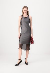 Schwarzes, transparentes Midikleid mit einer figurbetonten Silhouette, das einen runden Ausschnitt und ein Logo-Detail aufweist. Tragen Sie es mit schwarzen High Heels und einer kleinen roten Handtasche.