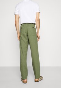 Mand iført olivengrønne højtaljede bukser med knappede baglommer, en hvid t-shirt og beige slid-sandaler, som står op imod en hvid baggrund.