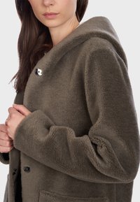 Cappotto con cappuccio in fleece verde oliva, con superficie testurizzata, ampie tasche frontali e dettagli in bottoni neri. Le maniche sono leggermente a sbuffo.