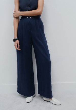 Kvinde iført navyblå ærmeløs jumpsuit med brede ben og hvide loafers, stående mod en ensfarvet lys væg.