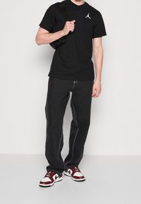 T-shirt nera con il logo Jordan bianco, abbinata a jeans scuri con cuciture bianche e sneakers basse rosse e bianche.
