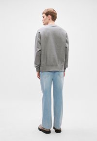 Šedá mikina s kleslými rameny, žebrovými manžetami a lemem, kombinovaná s světle modrými širokými džíny a černými botami. Modelka je otočená zády.