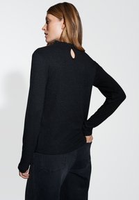 Schwarzes Langarmshirt mit einer strukturierten, funkelnden Oberfläche, mit einem Rüschensaum und einem Schlüsselloch-Ausschnitt auf der Rückseite. Kombiniert mit dunklen Hosen.