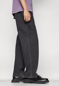 Pantalones de mezclilla negros de pierna ancha con una textura ligeramente descolorida, combinados con zapatos negros de suela gruesa y una camiseta morada de corte holgado.