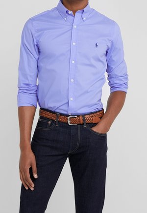 Man wearing a light blue button-up shirt with rolled sleeves, dark jeans, and a brown woven leather belt, standing with one hand in pocket.