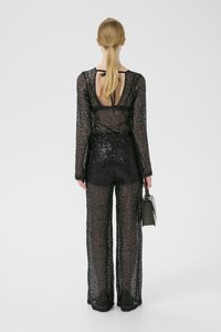 Sort gennemsigtig jumpsuit med lange ærmer, glitrende palietter, åben ryg og bindelukning. Modellen holder en lille grå håndtaske.