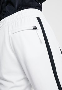 Vita sportbyxor med en zipficka, svarta sidostriper och elastisk midja. Slät textur med modern passform. Nike-logotyp synlig.