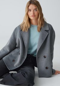 Grauer Wollmischung-Doppelt-Mantel mit abgerundeten Revers und braunen Knöpfen, getragen über einem hellblauen Hemd und schwarzen Hosen. Weicher Stoff.