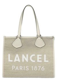 Sac beige avec le logo "LANCEL PARIS 1876" en blanc, fabriqué à partir de matériau tissé. Présente deux longues poignées et des accents en cuir sur les côtés.