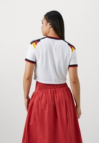 Vrouw met lang donker haar, gekleed in een wit crop top met zwarte, rode en gele strepen op de schouders en een rood rokje met hoge taille, staand met haar rug naar de kijker.