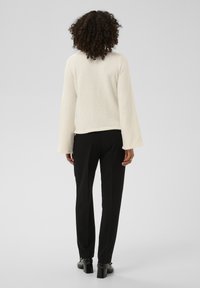 Pull-over crème texturé avec manches longues évasées ; assorti à un pantalon noir tailleur et des talons noirs épais.