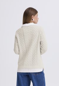 Light beige knitted sweater with textured cable patterns and ribbed hem, worn over a white collared shirt. Loose fit and long sleeves.