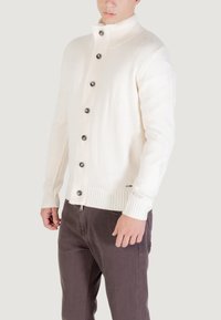 Hombre vestido con un suéter de punto color crema y pantalones marrón oscuro, de pie frente a un fondo claro.
