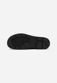 Semelle de chaussure noire présentant un motif de grip texturé, en caoutchouc épais, avec un design robuste aux contours définis et bords plats.