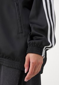 Chaqueta deportiva negra hecha de un material suave y ligero, con rayas blancas en las mangas, puños elásticos y un bolsillo con cremallera.