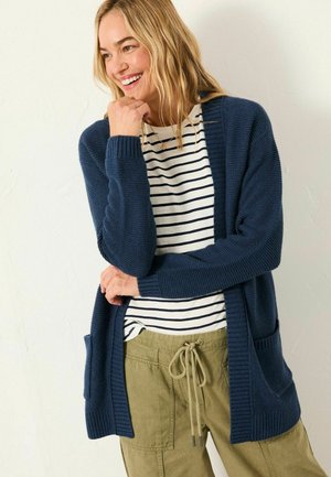 Marineblauer Strickcardigan mit offenem Vorderteil, gerippten Bündchen und zwei Fronttaschen, getragen über einem weiß-und-marinen gestreiften Hemd, kombiniert mit olivgrünen Hosen.