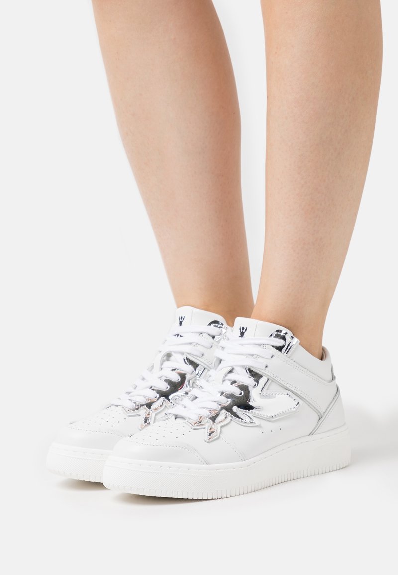 Baskets montantes blanches avec des détails d'étoiles réfléchissantes argentées portées sur des jambes nues sur fond blanc.
