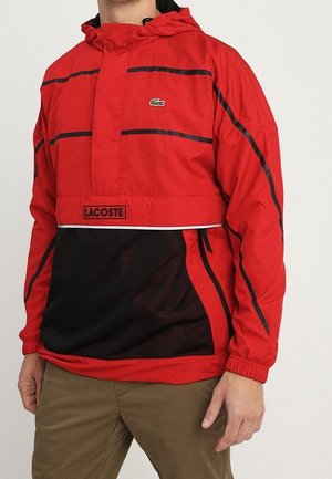 Roter und schwarzer Lacoste Kapuzen-Windbreaker mit vorderer Mesh-Tasche und schwarzen Streifendetails, getragen mit braunen Hosen.