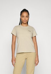 Mulher com cabelo trançado a usar uma t-shirt bege da Patagonia e calças cáqui, de pé contra um fundo cinza claro liso.