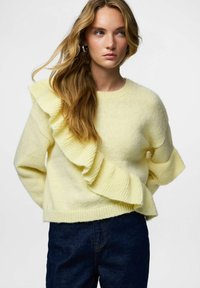 Żółty dziergany sweter z wyraźną falbaną po lewej stronie. Miękka faktura, dekolt w serek i ściągacze na mankietach. Noszony z ciemnymi dżinsami.