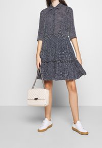 Marinblå prickig klänning med krage, trekvartslånga ärmar och flikig volang nederkant; ihop med en vadderad gräddfärgad handväska och vita sneakers.