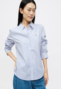 Modrá a biela pruhovaná košeľa na gombíky s klasickým golierom, rukávmi s možnosťou rolovania, prednou vreckom a malým detailom loga na hrudi.