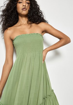 Kobieta o kręconych włosach, ubrana w zieloną sukienkę bez ramiączek, pozuje z jedną ręką na biodrze na jednolitym tle.