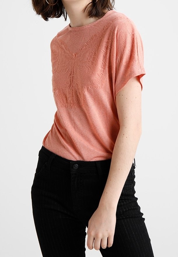 Femme portant un haut en dentelle corail à manches courtes rentré dans un pantalon noir taille haute avec de fines rayures, debout devant un fond uni.