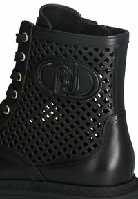 Black ankle boot with a perforated design, leather and synthetic upper, gold eyelets, and textured sole. Logo embossed on the side.